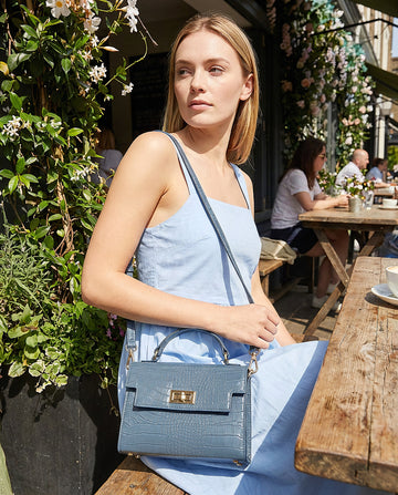 Woman with the Dusty Blue Margot Croc crossbody bag at a sunlit café terrace