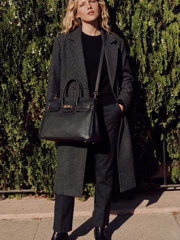 Woman in a grey wool coat wearing the Black Letizia Calfskin Bag by Clemés on her shoulder with the detachable strap, in front of a green hedge