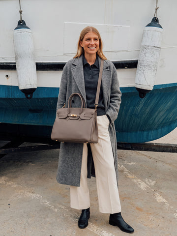 Woman in a grey coat and cream trousers wearing the Taupe Letizia Calfskin Bag by Clemés on the shoulder strap beside a harbour boat