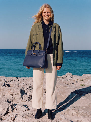 Woman in an olive jacket and cream trousers wearing the Navy Letizia Calfskin Bag by Clemés on the detachable shoulder strap by the sea