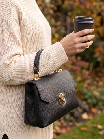 Woman with the Black Lola Dollaro crossbody bag on the top handle at an autumn garden table with coffee