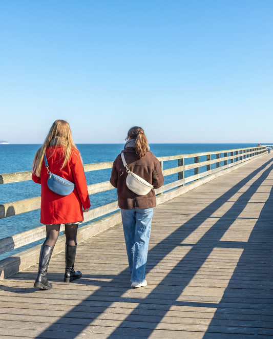 Romy Genuine Leather Crossbody Bum Bag in Denim Blue and Ivory, worn by two women walking along a seaside pier in summer