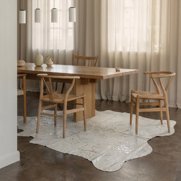 Silver metallic cowhide rug styled under an oak dining table on a dark concrete floor
