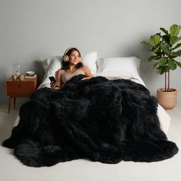 Black Octo New Zealand sheepskin rug throw styled on a bed, model relaxing with headphones and phone