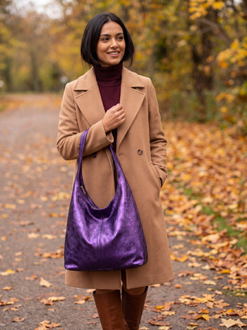 Woman carrying the Violet Silvia Metallic Suede Slouch Bag on an autumn tree-lined path with fallen leaves