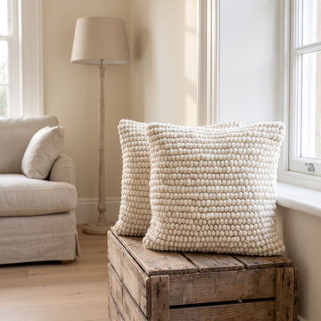 Two natural ivory felted wool square cushions on a rustic wooden crate beside a sash window in a cream living room