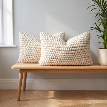 Two natural ivory felted wool lumbar cushions on an oak bench against a pale grey wall with a green plant