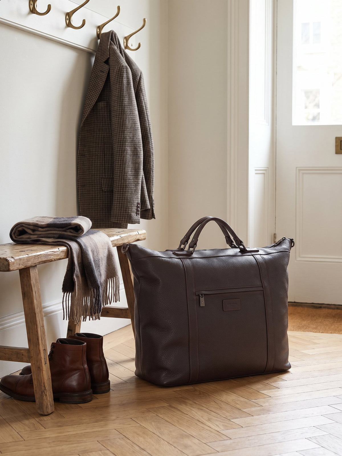 Alessio dark brown leather travel bag by Clemés in a townhouse hallway beside a bench and boots
