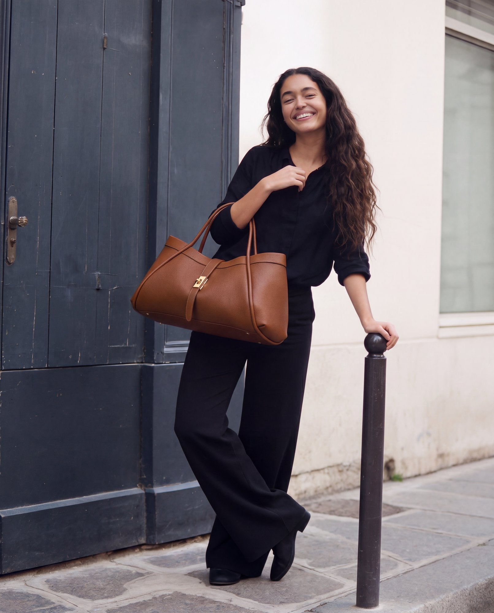 Woman with the Dark Tan Bianca Large Leather Handbag by Clemés beside an iron bollard on a cobblestone street