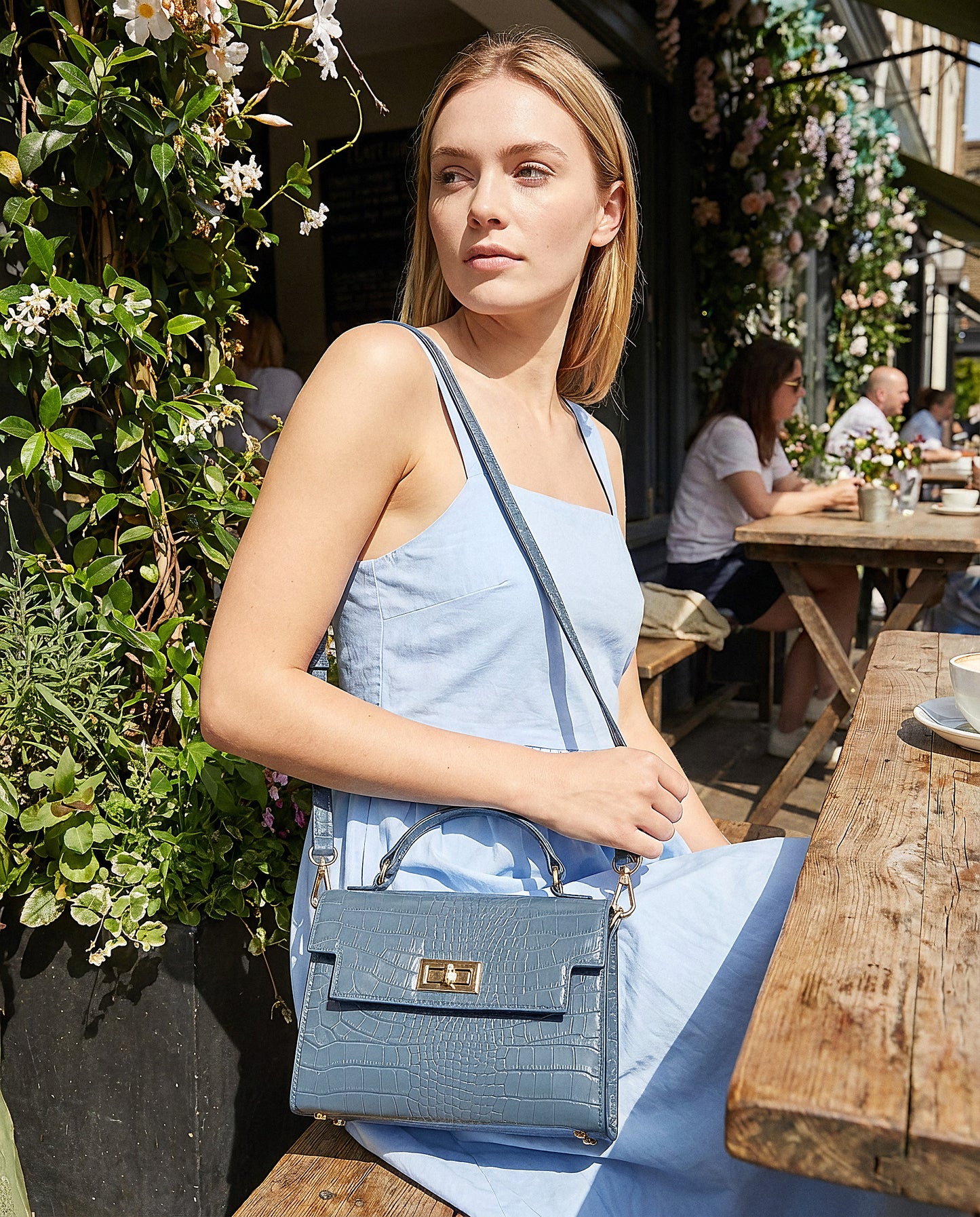 Woman with the Dusty Blue Margot Croc crossbody bag at a sunlit café terrace