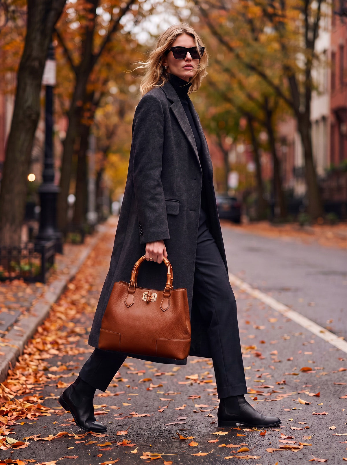 Woman in a charcoal wool coat walking along an autumn street carrying the Ginevra Bamboo-Handle Leather Bag by Clemés in Cognac