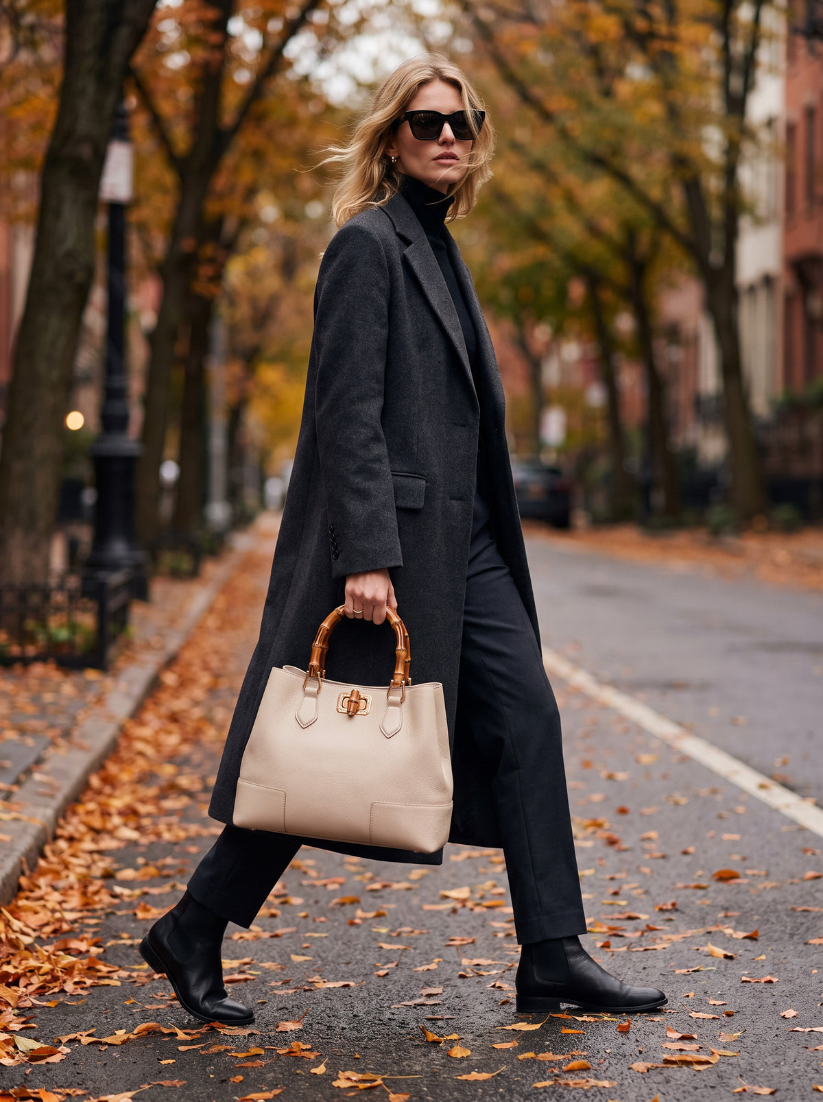 Woman in a charcoal wool coat walking along an autumn street carrying the Ginevra Bamboo-Handle Leather Bag by Clemés in Oyster
