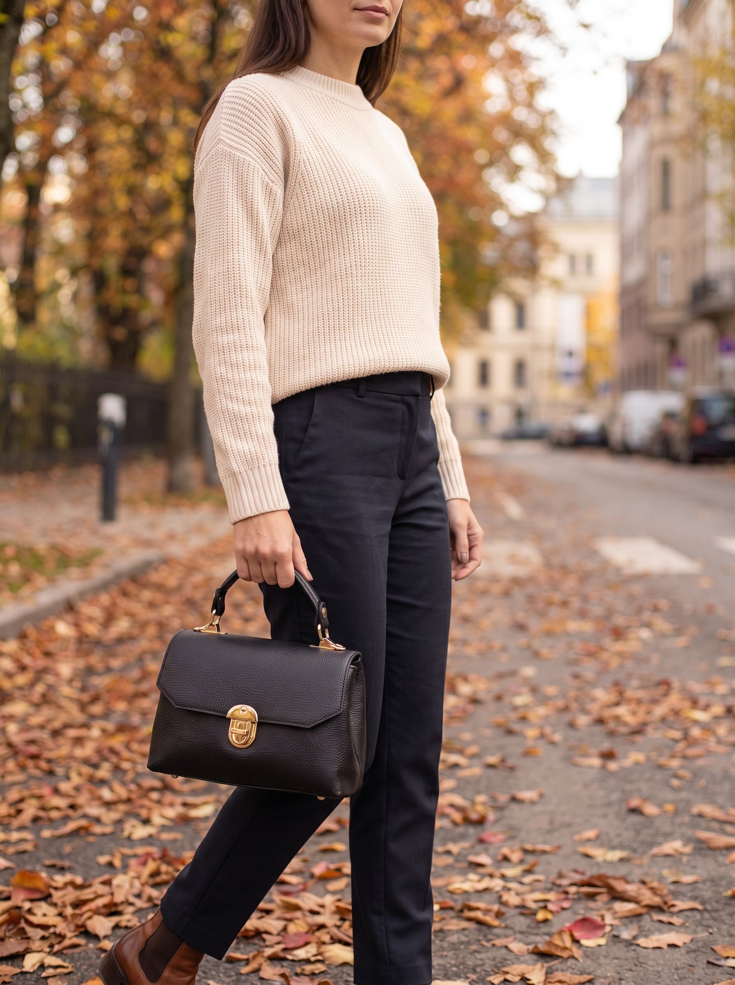 Woman carrying the Black Lola Dollaro crossbody bag on the top handle while walking on an autumn city street