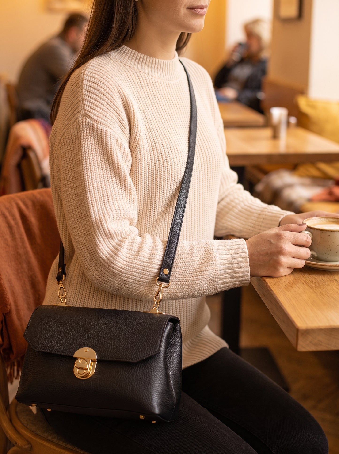 Woman wearing the Black Lola Dollaro bag crossbody on its adjustable shoulder strap at a cosy cafe table