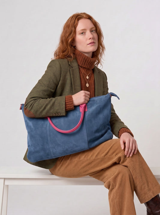Model seated on a white bench carrying the Blue Milano Max Suede Bag by Clemés on her shoulder, fuchsia pink handle