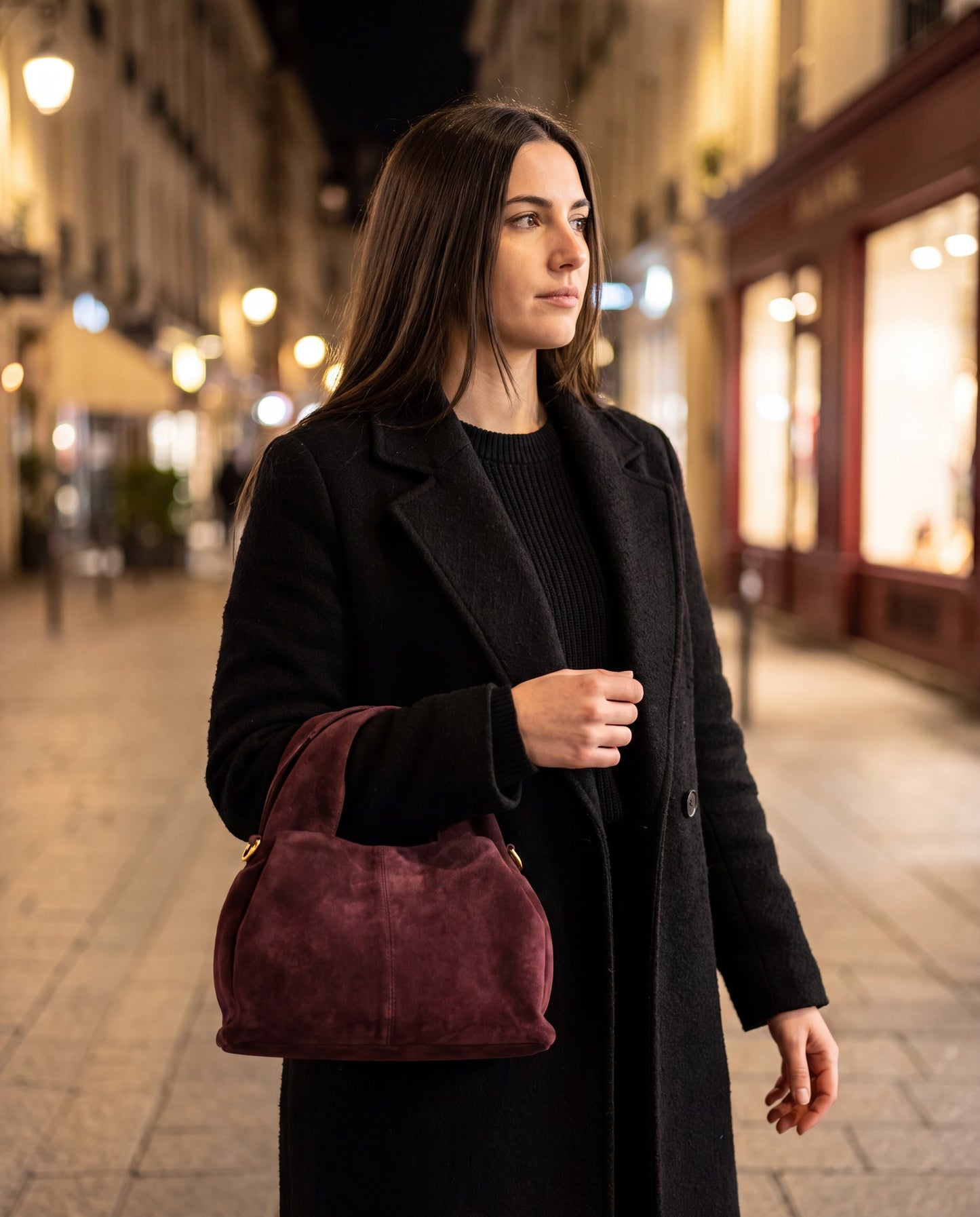 Woman carrying the Burgundy Noemi Suede Handbag on a city street lit by evening shop lights