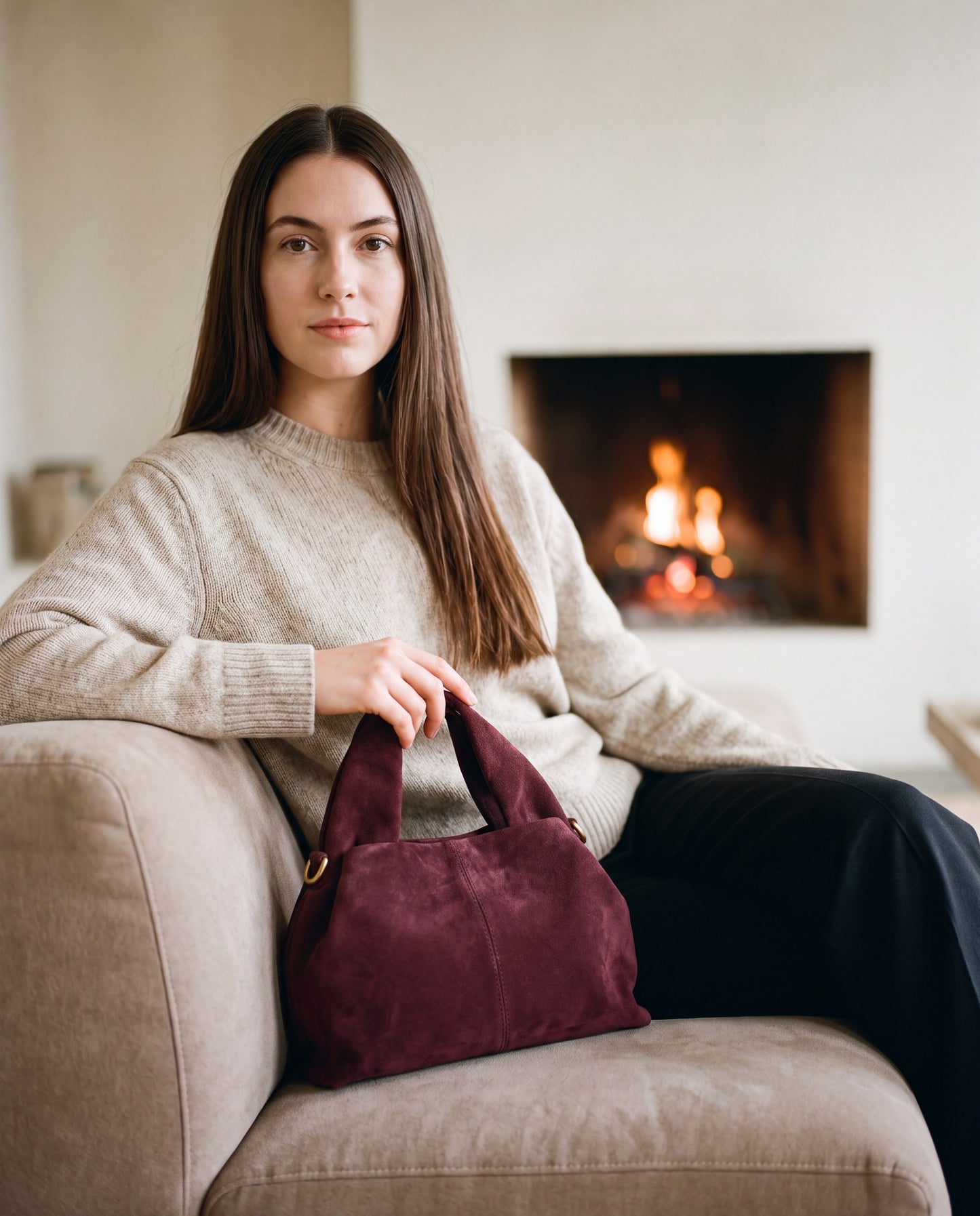 Woman seated by a fireplace with the Burgundy Noemi Suede Handbag beside her