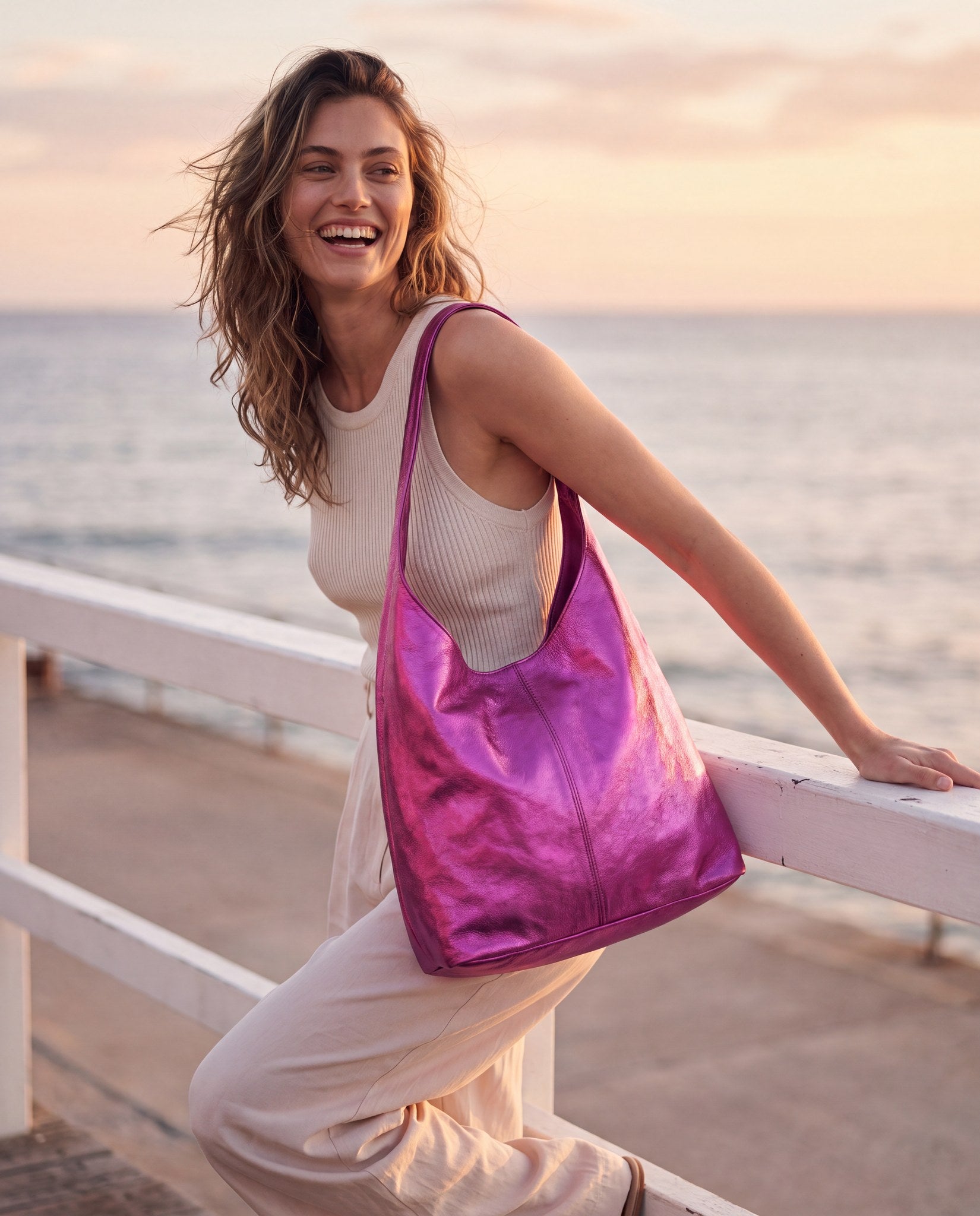 Woman with the Fuchsia Silvia Metallic Suede Slouch Bag on a seaside promenade at golden hour
