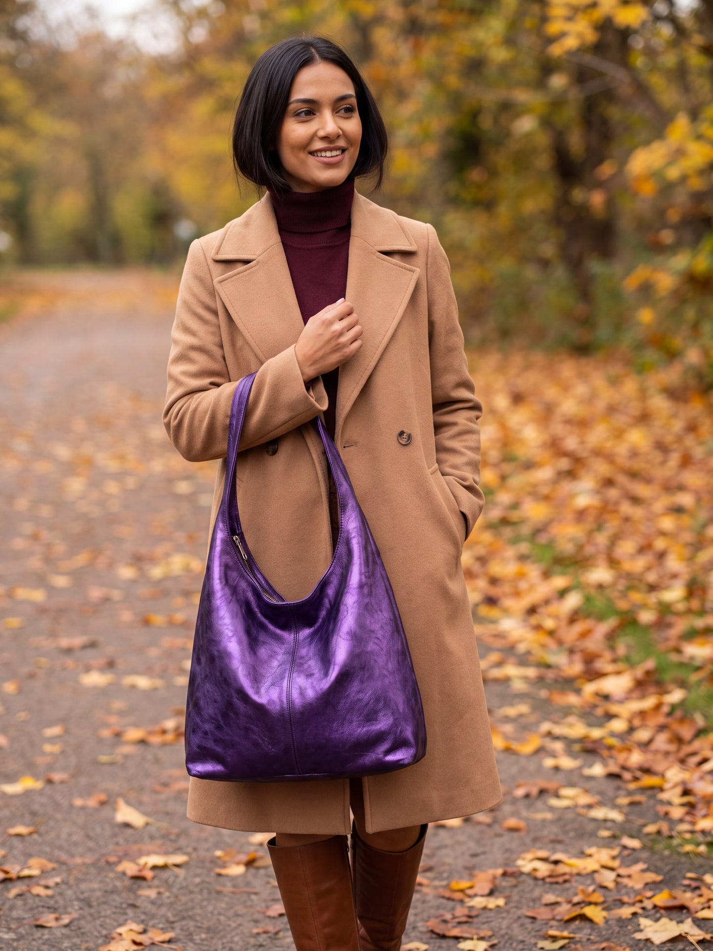 Woman carrying the Violet Silvia Metallic Suede Slouch Bag on an autumn tree-lined path with fallen leaves
