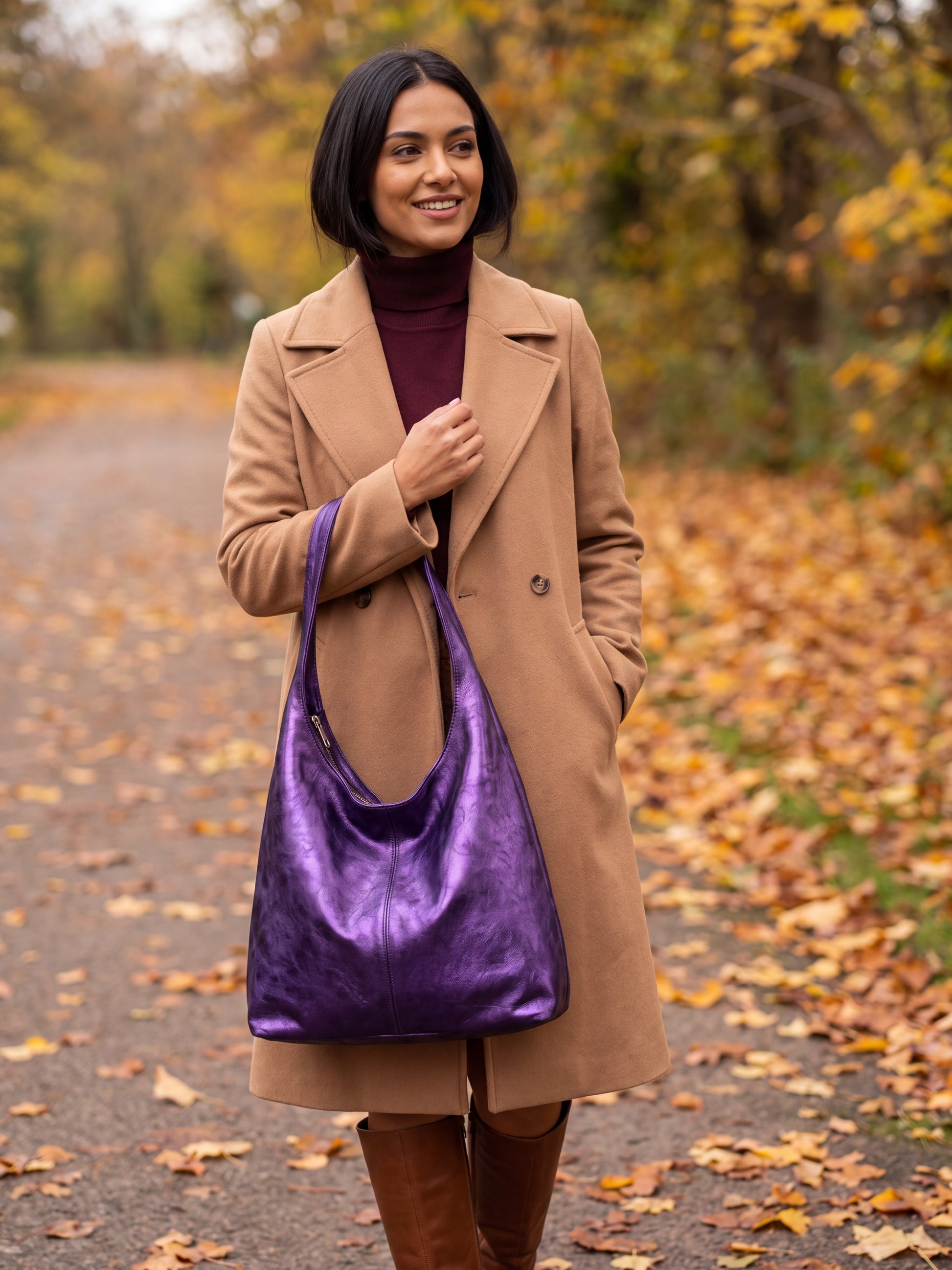 Woman carrying the Violet Silvia Metallic Suede Slouch Bag on an autumn tree-lined path with fallen leaves